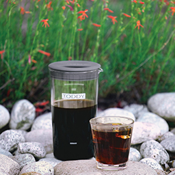 Toddy Artisan small batch cold brewer with coffee and glass of cold brew coffee on rocks outside by flowers